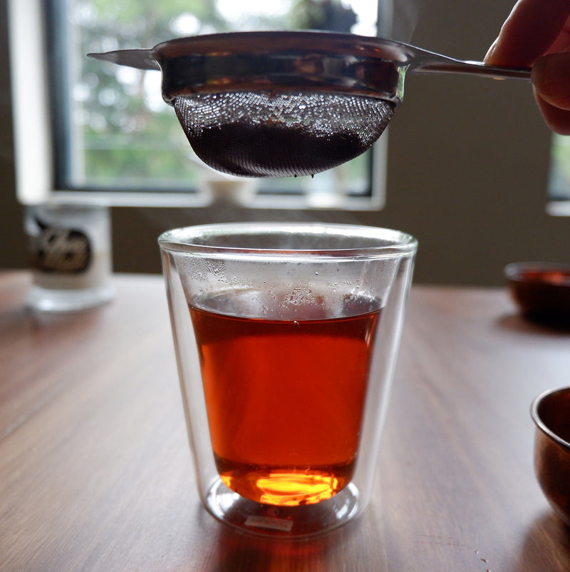 A glass with  Chai Walli English Breakfast black tea and a hand holding a strainer above it