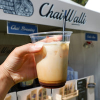 A hand holding an glass with ice tea made with chai syrup in it with the Chai Walli banner in the backdrop