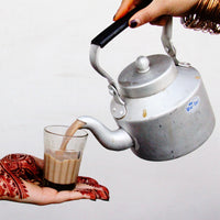 a henna decorated hand holding a chai glass while another hand pours chai in it from a kettle